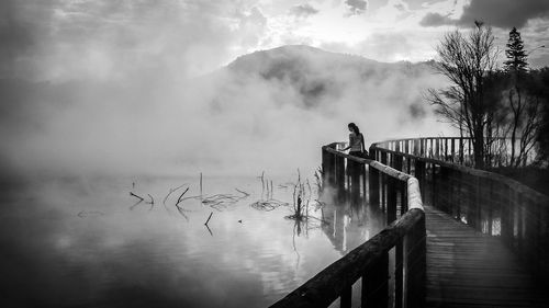 Rear view of woman standing on wooden walkway by lake during foggy weather