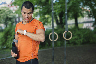 Man checking smart watch and holding bottle while standing at park