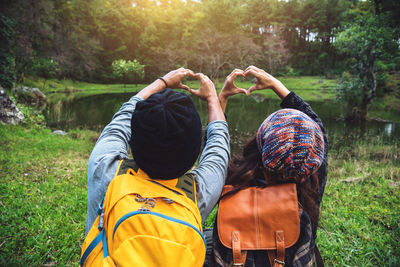 Rear view of couple making heart shape while sitting by lake at forest