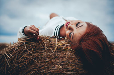 Young woman lying on hay bale against cloudy sky at farm