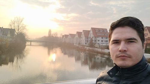 Portrait of young man standing by river in city against sky during sunset