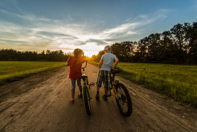 Rear view of friends standing with bicycles on dirt road against sky during sunset