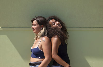 Side view of two young women standing against green wall