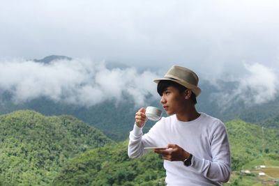 Young man having coffee while sitting against mountain during foggy weather