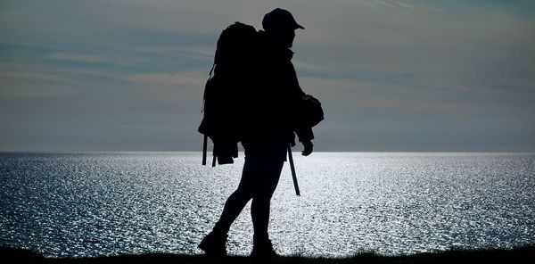 Silhouette woman carrying backpack while standing at beach against sky