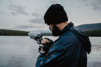 Side view of serious man in warm clothes reviewing photos on camera standing on beach of lake on cloudy day