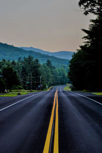 Empty road along trees and mountains against sky