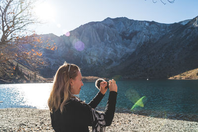 Blonde haired woman at beautiful mountain lake filming with video camera as sun begins to set 