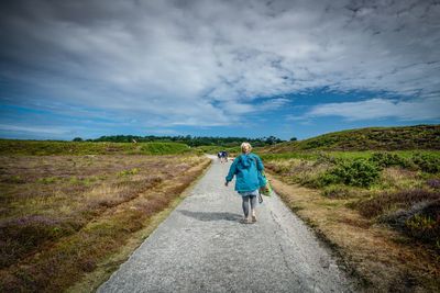 Rear view of senior woman walking on road amidst landscape against cloudy sky