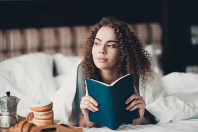 Contemplative woman with book lying on bed at home