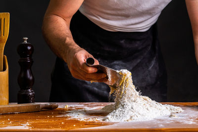 Crop man mixing wheat flour with egg using scraper at wooden table in bakery