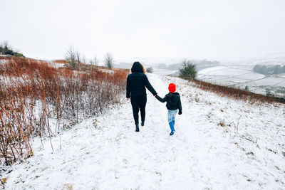Rear view of mother and son holding hands while walking on snow covered land