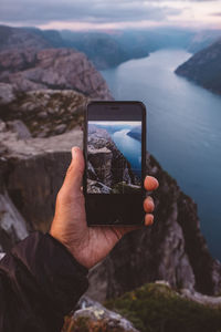 Hand holding smartphone with image of scene in background on it at norwegian fjords.