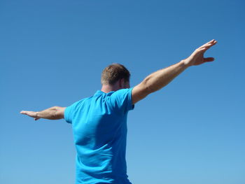 Low angle view of man standing with arms outstretched against clear blue sky