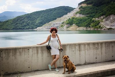 Smiling pregnant woman with dog standing on bridge over lake against mountains