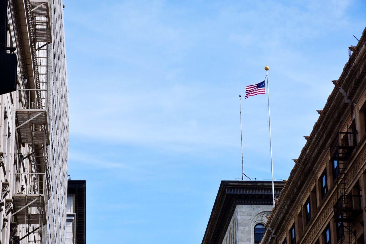 building exterior, architecture, low angle view, built structure, flag, national flag, patriotism, identity, american flag, sky, city, culture, day, building, pride, tower, tall - high, outdoors, wind, cloud - sky
