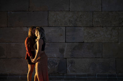 Two girls standing against stone wall during sunset