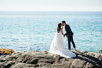 Newlywed couple kissing while standing at rocky beach against clear sky