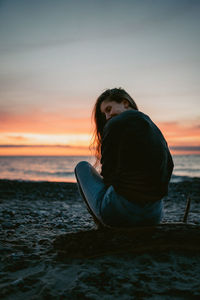 Woman sitting on shore at beach against sky during sunset