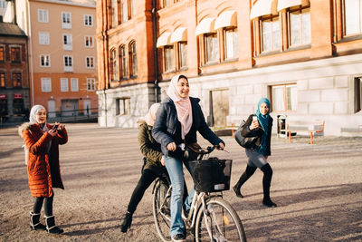 Young women photographing teenage girl cycling with friend against building in city