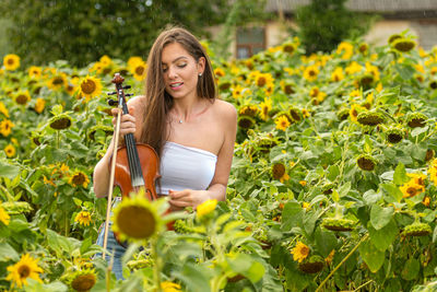 Young slim brunette with long hair standing in sunflower field and playing violin in rainy day