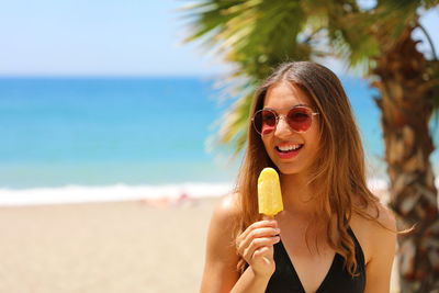 Young woman in sunglasses eating popsicle while standing at beach during sunny day
