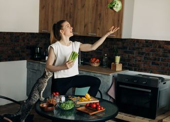 Cheerful young woman preparing green salad at home and jokingly throwing vegetables up