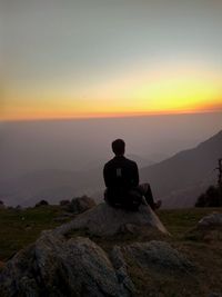Rear view of man sitting on rock against sky during sunset