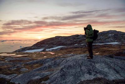 Full length of hiker photographing while standing on rock against sky during sunset