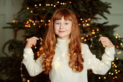 Happy young girl sitting by the christmas tree with garland