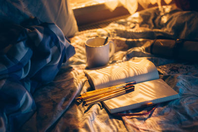 Coffee cup by book and pencils on bed at home