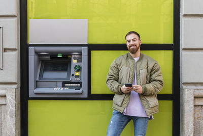 Smiling young man with smart phone standing by atm