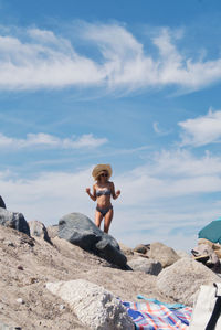 Full length of shirtless woman with bikini on rock at beach against sky