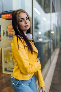 Side view of calm young female in trendy bright yellow leather jacket and jeans standing near glass wall of modern city building and looking at camera