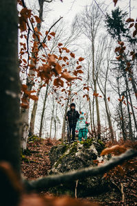 Low angle view of girls standing amidst bare trees in forest during autumn