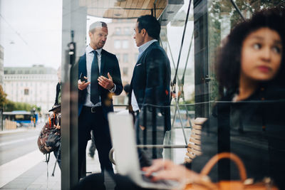 Mature male business coworkers talking while standing at bus stop seen from glass