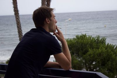 Young man smoking at balcony against sea