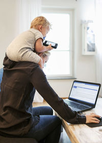 Mother using laptop while carrying baby boy on shoulders at home