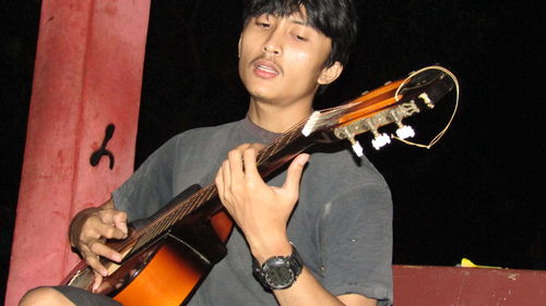 Close-up of young man playing guitar while sitting outdoors at night