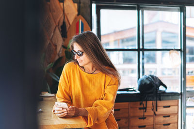 Young woman with long blonde hair wearing sunglasses and yellow longsleeve with mobile phone in cafe