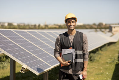 Portrait of smiling male engineer holding documents and digital tablet while standing near solar panels at power station