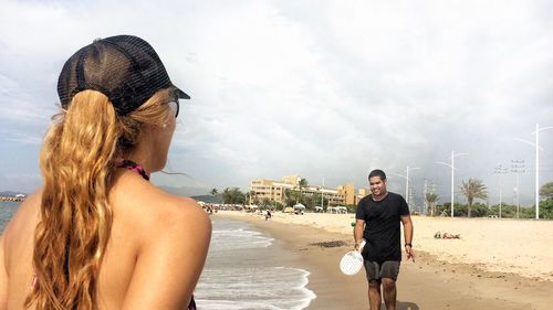 Woman with long hair looking at friend playing table tennis at beach against sky