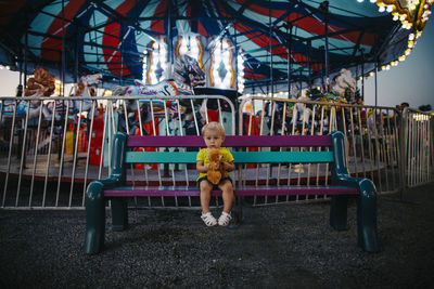 Portrait of baby girl holding toy while sitting on bench at amusement park