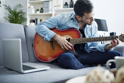 Young man at home sitting on couch playing guitar next to laptop