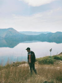 Boy standing on grass by lake against mountains and sky