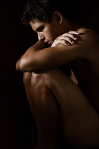 Shirtless lonely young man sitting against black background