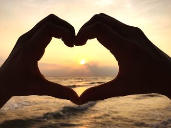 Cropped image of hands making heart shape against sea during sunset