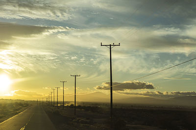 Road by electricity pylon against sky during sunset