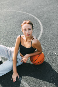 Portrait of a charming girl sitting on a sports field in a park or school with a basketball 