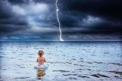 Rear view of shirtless girl with inflatable ring standing in sea against cloudy sky